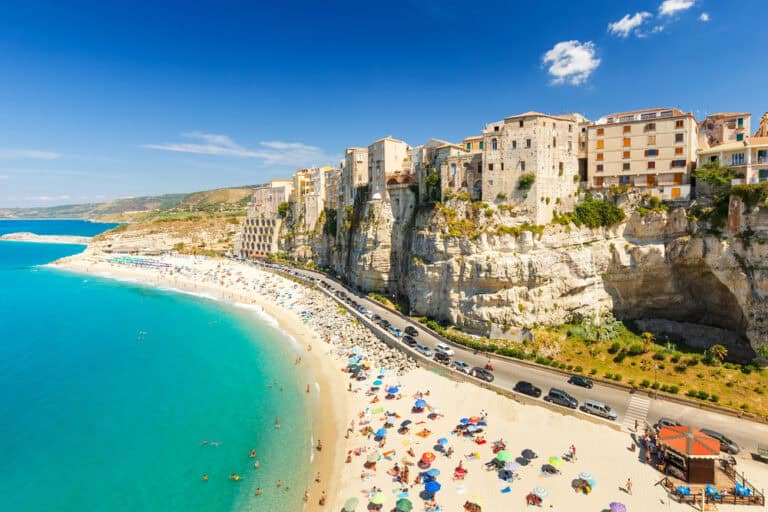 aerial image, historic buildings of tropea on the edge of the white cliffs, a genly winding road at the base of the cliffs and the white sand beach and blue sea on a sunny day.