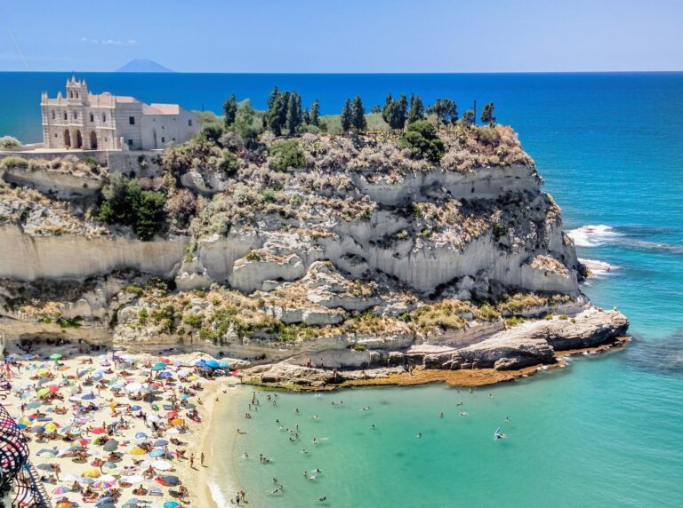 Historic church of Santa Maria della Isola in Tropea on rocky outcrop with white sand beach and clear blue sea below on a sunny day. Colourful beach umbrellas and people swimming in the sea.